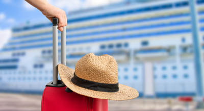 A person leaning on a red, wheeled sutcase in front of a docked cruise ship.